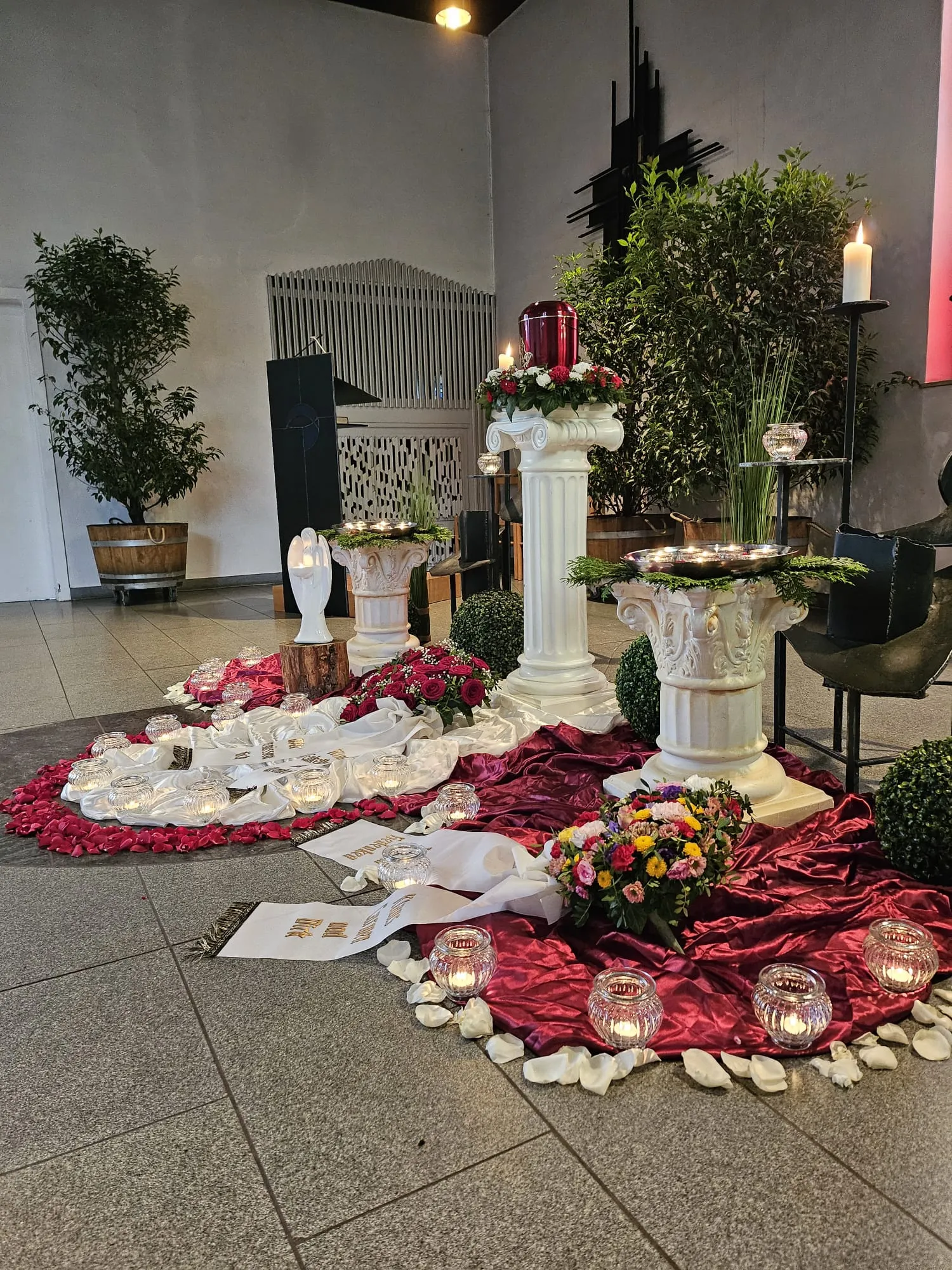 Festlich gestaltete Trauerfeier mit roter Urne, Säulen und Blumenschmuck