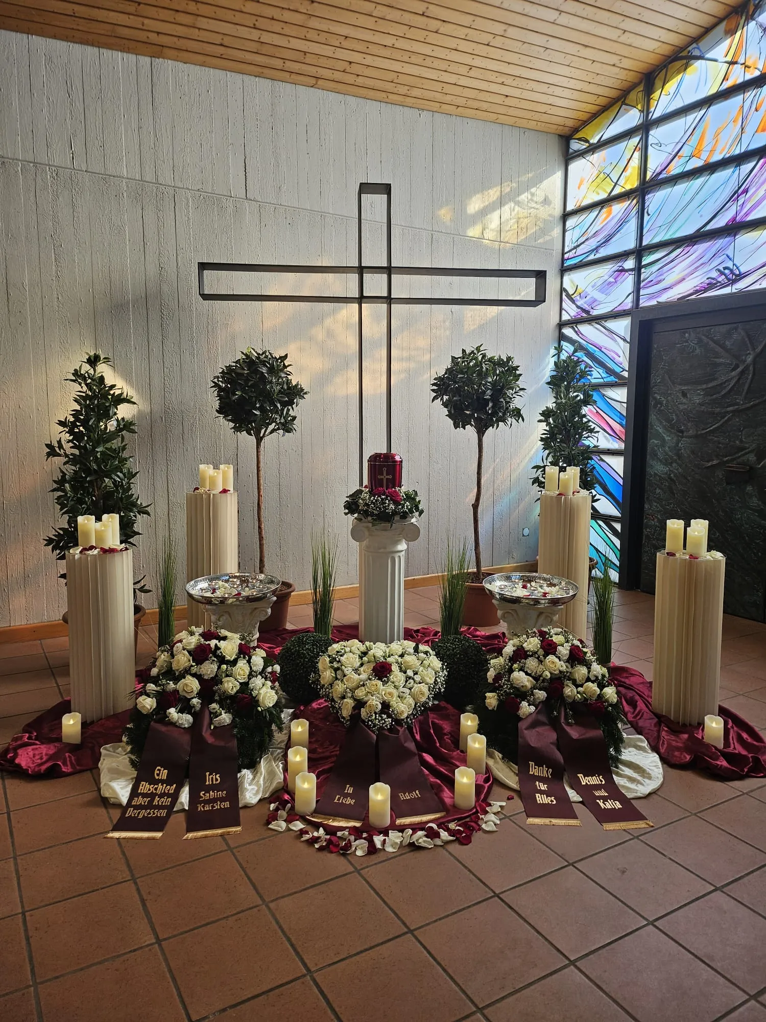 Trauerfeier mit roter Urne auf Säule und Blumenherzen mit Trauerschleifen