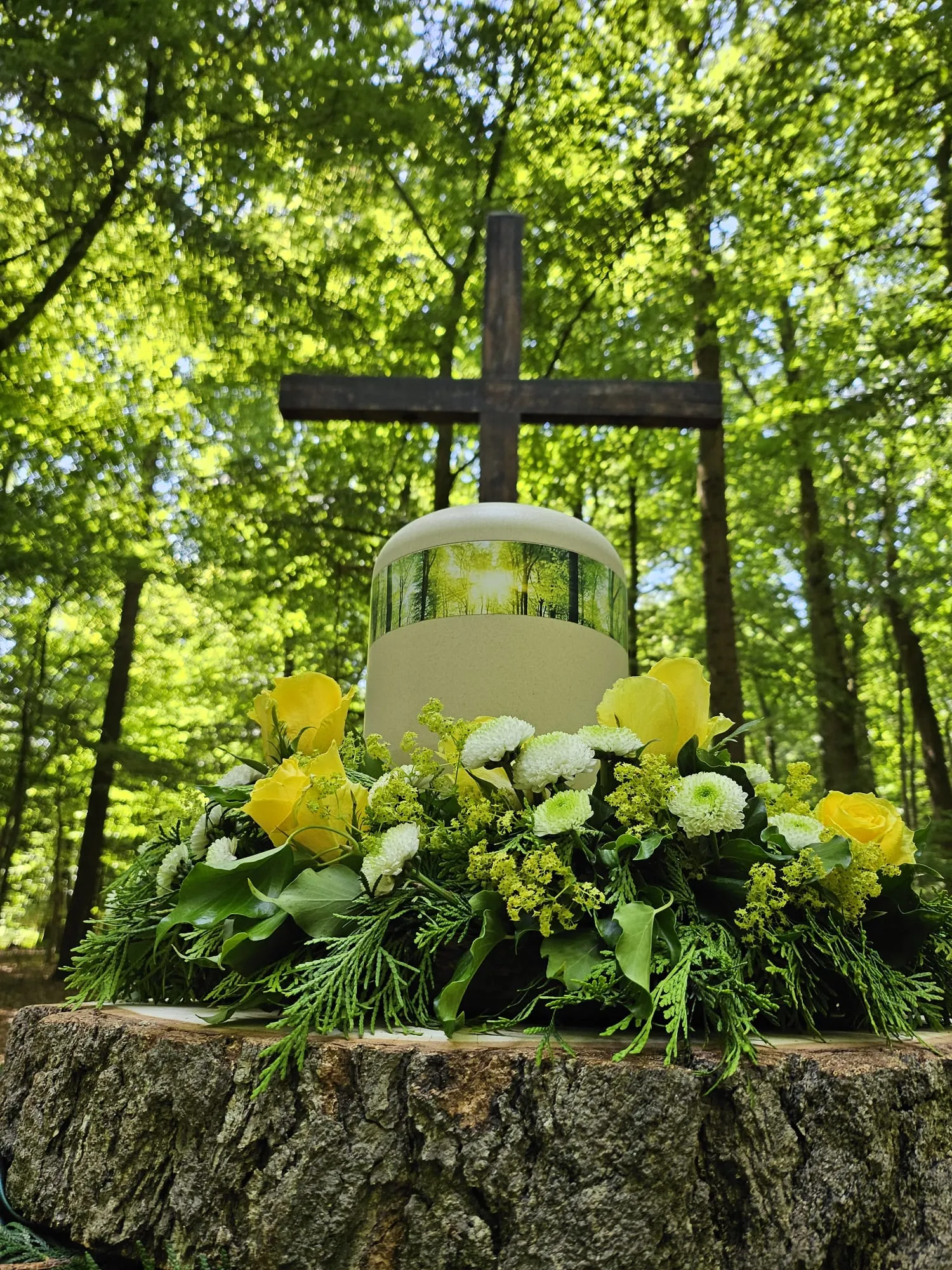 Naturabbaubare Urne mit Waldmotiv und gelben Rosen vor einem Holzkreuz im Wald